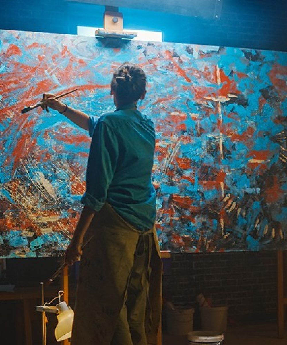 Talented Female Artist Works on Abstract Oil Painting, Using Paint Brush She Creates Modern Masterpiece. Dark and Messy Creative Studio where Large Canvas Stands on Easel Illuminated.